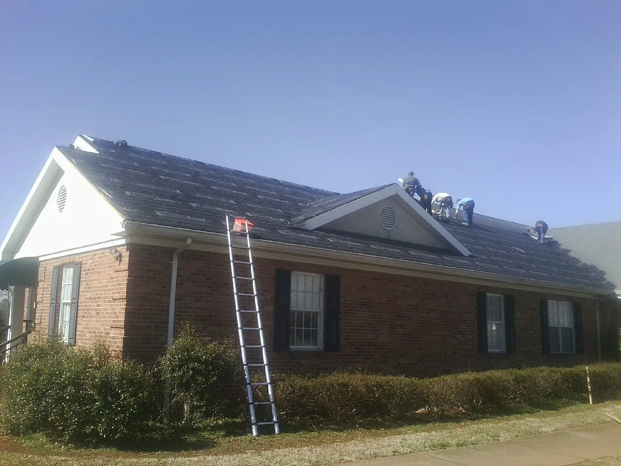 Crew tearing shingles off a brick ranch roof with a ladder set — Emergency Roof Repair work by our crew in Lanark