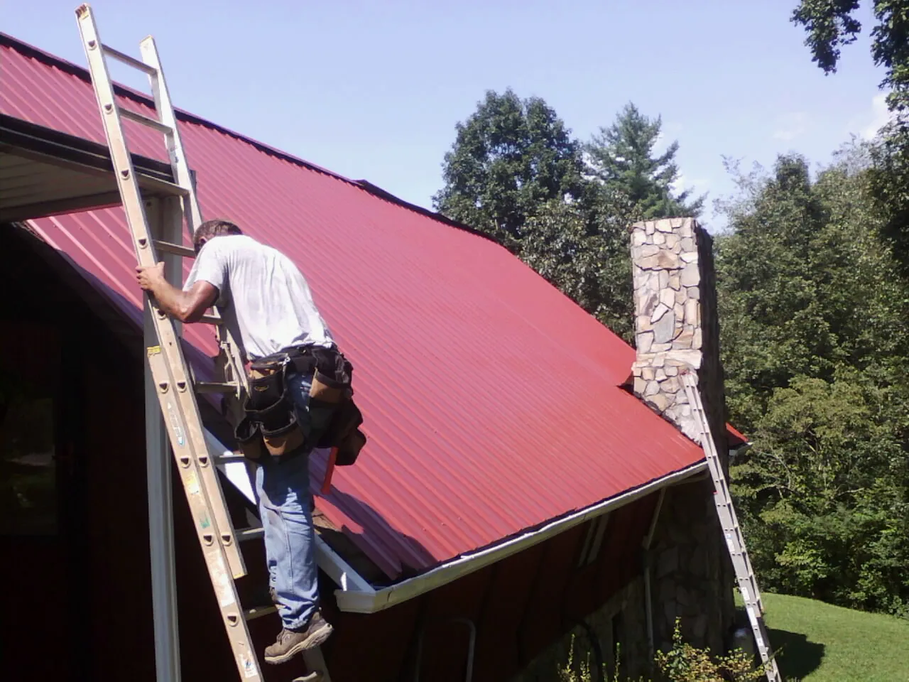 Roofer on a ladder beside a red metal roof and stone chimney — Soffit & Fascia Repair by Roofing Brothers in Lanark
